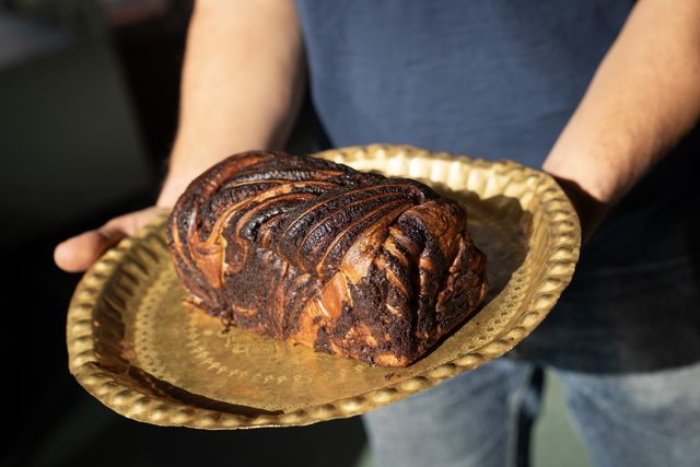 A person holds a golden textured tray with a loaf of dark, swirled chocolate bread or cake. The person wears a blue shirt and jeans.